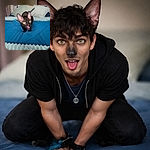 A quirky black cat with enormous ears and bright yellow eyes is caught mid-lick on a cozy blue bedspread, surrounded by soft pillows and homey decor, looking both curious and mischievous.