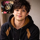 A fluffy black and tan dog with big, expressive eyes looks up curiously while sitting among a cozy pile of blankets and a pink slipper on a wooden floor, surrounded by household clutter.