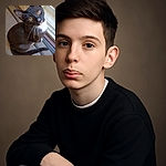 A curious tuxedo cat with bright amber eyes looks up with an inquisitive tilt, sitting on a textured rug near a glass door, ready to pounce on the next adventure or maybe just a sunbeam.