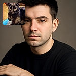A sleek black cat is perched comfortably on someone's lap, its shiny fur reflecting the warm indoor light. The background shows a cozy room with wooden furniture and a carpeted floor, while the cat's intense gaze adds a touch of mystery and charm.