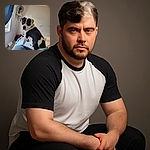 A charming black and white dog sits on a bed, gazing back with soulful eyes. The cozy bedroom features a fan on a wall shelf, soft natural light from a window, and a comfy blanket in the foreground, creating a warm and relaxed atmosphere.