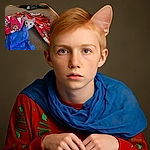 A curious orange tabby cat peeks out wide-eyed from beneath a pile of colorful Christmas wrapping paper and blue tissue paper, looking like it just found the best hiding spot ever during holiday chaos.