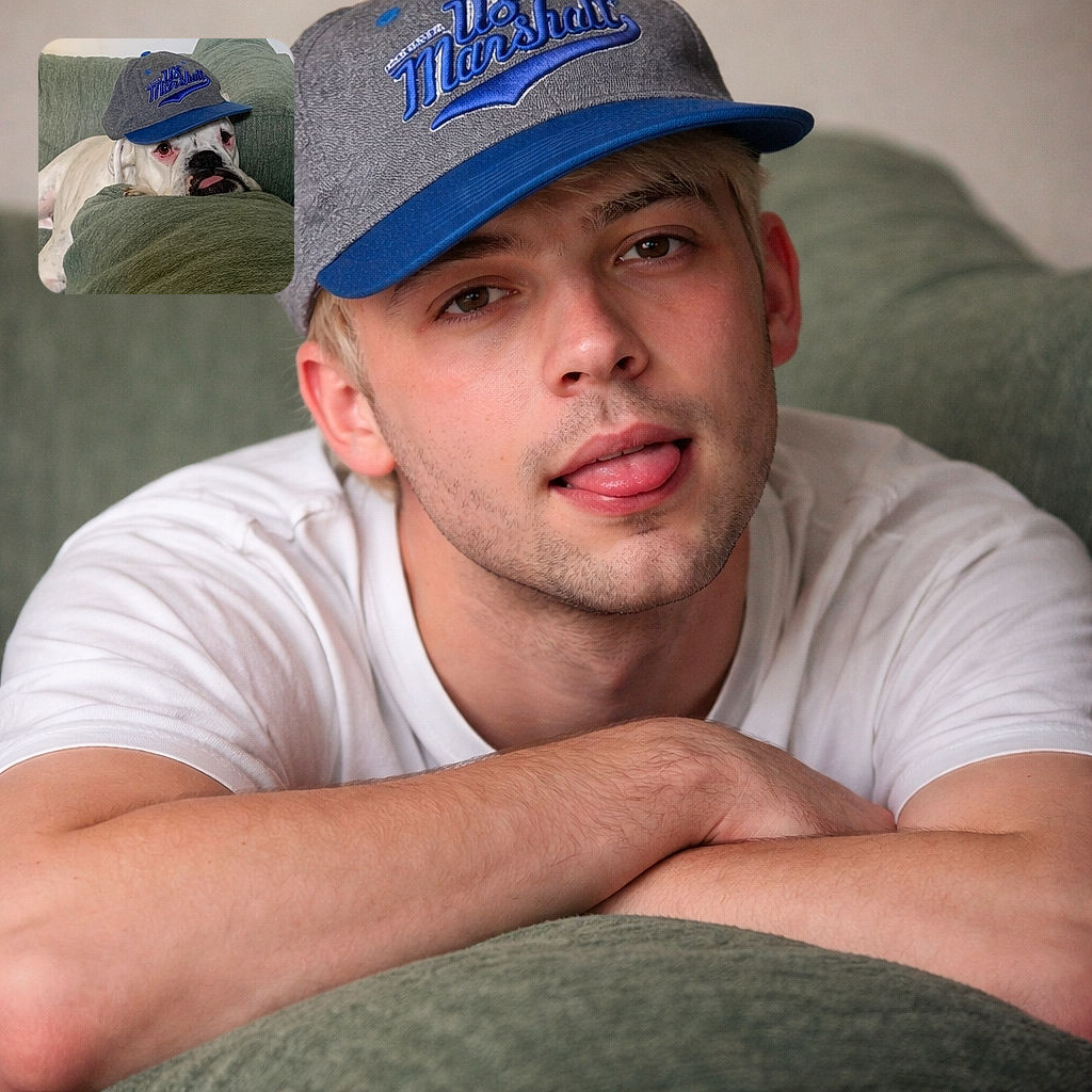 A weary white boxer lounges on a green couch wearing a slightly oversized baseball cap, tongue peeking out as if the hat exhausted him more than the day did. The textured cushions form a cozy fortress around his head while the cap's logo adds accidental swagger to this nap-time portrait.