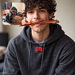 A proud, curly-haired dog sits on a bed holding a long bacon strip like a championship trophy, fur obscuring most of its eyes and a red tag bobbing on its collar. The cozy bedroom behind it has a poster on the wall, a TV, a cluttered desk with a mirror and toiletries, and a backpack — the dog clearly staged this heist and demands applause (and maybe another treat).