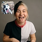 A happy black and white dog sitting on a rocky ground with its tongue out, looking up at the camera with a joyful expression. In the background, a person's legs and shoes are visible, standing on the same rocky terrain.