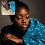 A cozy black dog snugly wrapped in a blue blanket with white patterns, peacefully napping on a textured cushion. The warm, homely setting invites a sense of calm and comfort.