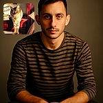 A regal tabby cat basks in the warm glow of sunlight while perched elegantly on a wooden dresser drawer, casting a serious yet curious gaze at the camera. The cozy bedroom setting with soft shadows and a framed picture adds a homey charm.