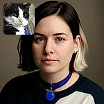 A close-up of a black and white cat with a slightly blurred focus, showing its calm and curious expression, with a blue tag visible on its collar and a cozy indoor background.