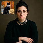 A dignified black and white cat sits gracefully on a stone path surrounded by pebbles, with a wooden fence and brick wall in the background. The cat wears a collar with a bell and a round tag, looking alert and regal.