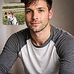 A curious and attentive dog with a brown, black, and white coat sits on a grassy field near a calm river, with a scenic countryside backdrop featuring trees and houses in the distance under soft daylight.