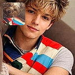 A charming small dog sporting a colorful striped shirt lounges comfortably on a couch, giving the camera a curious and slightly tilted head look, as if ready for its close-up or a treat.