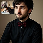 A sleek black cat with bright green eyes and a cute collar poses curiously on a cozy blue blanket, with a playful scratching post and colorful toys scattered on a soft carpet in a warm, inviting living room.