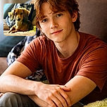 A cute brown dog lounges comfortably on a couch with a soft green blanket, looking attentively at the camera while surrounded by cozy household items and a patterned jacket in the background.