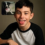 A playful black and white puppy with its tongue sticking out, sitting on a couch with a pillow that reads 'Aloe Vera Bamboo' in the background. The image is a bit blurry and dimly lit, giving it a cozy, casual snapshot vibe.