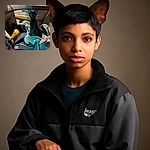 A sleek black kitten with big ears sits alertly on a pile of jackets inside a car, looking curiously at the camera. The warm interior lighting contrasts with the dark night outside the window, and the wooden gear shift adds a vintage vibe to the cozy scene.