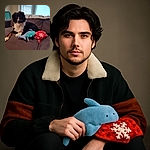 A fluffy black and white dog lounges comfortably on a gray couch, surrounded by a cozy red snowflake blanket and a plush blue shark toy, with soft window light illuminating the scene.