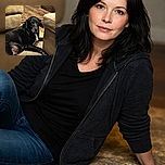 A relaxed black dog with a white patch on its chest lounges comfortably on a patterned rug between two beige couches in a cozy living room with wooden floors and a rustic chest in the background.