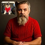 A charming schnauzer dog wearing a cozy red sweater sits proudly on a black leather couch, looking slightly to the side with a thoughtful expression. The background is softly blurred, emphasizing the dog's fluffy beard and expressive eyes.