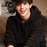 A fluffy black Pomeranian dog sits happily on a bed with a patterned blanket, tongue out and eyes wide, in a cozy room with a TV and various furniture in the background.