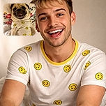A cheerful pug wearing a white shirt adorned with yellow smiley faces, lounging comfortably on a floral-patterned pillow and white bedding, flashing a toothy grin that could brighten anyone's day.