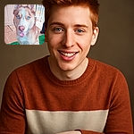 A close-up photo of a happy husky with striking bright blue eyes and a pink nose, sitting on a green mat with a wooden wall and railing in the background. The dog looks friendly and alert, tongue slightly out as if smiling.