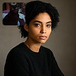 A mysterious black cat with glowing green eyes sits on a cozy bed, looking directly at the camera with an intense stare. The dimly lit room adds to the enigmatic vibe, with a slightly blurred background featuring a framed picture and some furniture.