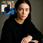 A sleek black cat with bright green eyes sits attentively on a blue blanket, posing like the mysterious ruler of the room. Behind it, a cozy bedroom scene unfolds with a wooden mirror, a leafy plant, and a vintage dresser, adding charm and warmth to the setting.