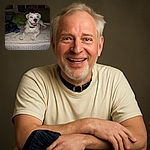 A happy white dog with a black nose and collar lounges on a mattress with a slightly worn cover, its eyes reflecting the camera flash like glowing orbs, surrounded by casual household items and a dimly lit background.