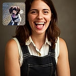 A happy dog with a shiny brown and white coat is sitting on a gravelly surface, looking up with its tongue out in a joyful expression, wearing a black harness and leash, basking in the sunlight.