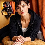 A dignified black dog lounges on a well-worn couch, giving the camera a wise, slightly judgmental stare — like it's silently calculating how many treats you'll admit to stealing. Warm orange curtains and cozy living-room tones frame the scene, highlighting the glossy fur, white chest patch, and expressive brown eyes.