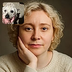A close-up shot of a scruffy white dog being gently held by a hand under its chin, with the dog's soulful eyes looking up. The background is blurry and nondescript, focusing all attention on the dog's adorable, slightly messy face.