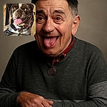 A happy pug with a brindle coat lies on a stone floor, tongue out and eyes sparkling with excitement, wearing a red collar with tags, looking like the ultimate good boy ready for some belly rubs.