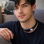 A close-up photo of a black cat with yellow eyes, wearing a collar, being gently held by a person's hand. The background shows a beige couch and a light blue wall, with soft lighting creating a cozy atmosphere.