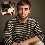A charming tabby and white cat lounges comfortably on a plush red blanket atop a black leather chair, gazing curiously at the camera with wide eyes in a cozy indoor setting.