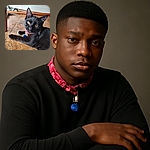 A sleek black cat with bright yellow eyes lounges on a textured floor, wearing a red collar with tags and a bell, looking curiously at the camera with a relaxed pose under a wooden chair.