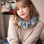 A curious cat with strikingly large blue eyes wears a floral scrunchie around its neck, perched on a cozy bed with a red blanket and a black hoodie in the foreground. The background shows a colorful poster and a TV, adding a casual home vibe to the scene.