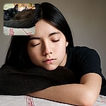 Two black cats are cozily curled up and sleeping on a fabric with red text, while a blurred shirtless person is in the background, out of focus. The cats' fur is soft and detailed, making them the clear stars of this sleepy snapshot.