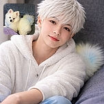 A fluffy white Pomeranian puppy lounges comfortably on a textured gray couch with a soft purple quilt in the foreground, looking adorably curious and slightly tilted head, radiating pure cuteness and cozy vibes.