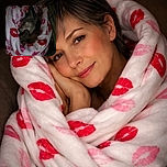 A cozy dog wrapped snugly in a blanket adorned with red and pink lip prints, peacefully sleeping in a dimly lit setting that adds a warm, intimate vibe despite the grainy quality.
