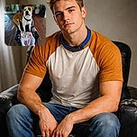 A charming dog with a blue collar stands proudly on a worn-out black chair with a cozy pink and white blanket draped over it. The wooden floor and homey background, including a spooky skull decoration and a framed dog picture, add character to this candid indoor scene.