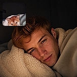 A sleepy red-and-white dog is tucked under a fluffy blanket, peeking out like a cozy little fox who has been caught mid-dream. The dim bedroom setting and snug bedding make this a wonderfully warm and drowsy portrait.