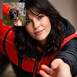A wet black dog wearing a bright red life jacket looks up with soulful eyes while reaching out to a human hand over murky water, capturing a moment of trust and companionship.