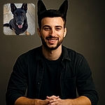 A happy black and tan dog with perky ears sits on a concrete floor inside a fenced area, looking up with a friendly smile and bright eyes, ready for some fun or a treat.