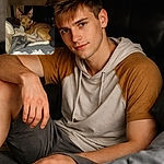 A small brown dog curled on a messy bed, giving the classic blue‑eye flash stare while surrounded by shredded tissues and scattered paper — caught mid-guilty nap with alert ears and a ‘who, me?’ expression.