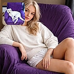 A relaxed white dog lounges comfortably on a purple-covered armchair, looking calmly at the camera with a serene expression. The setting appears to be indoors with a wooden floor and some visible household items in the background.