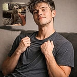 A fluffy gray cat sits upright on someone's lap like a tiny, judgmental king while a sleepy dog lounges on the couch behind it and a person's profile peeks in from the right — cozy living-room vibes and unbothered pets stealing the show.