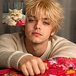 A grumpy little floof lounges like tiny royalty — one paw stretched toward a colorful blanket, half-lidded eyes judging your life choices while a soft curtain bokeh provides the dramatic backdrop.