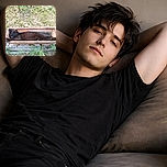 A black cat is cozily stretched out inside a narrow rectangular planter outdoors, blending in with the earthy tones and surrounded by grass and small plants, perfectly demonstrating the art of fitting into unexpected spots for a nap.