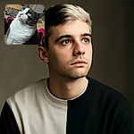 A close-up shot of a curious tuxedo cat with striking green eyes gazing upward, surrounded by a cozy indoor setting featuring scattered clothes and vibrant pink slippers in the background.