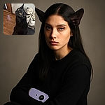 A sleek black cat with striking yellow eyes looks up with a curious expression while sitting on a black surface next to a purple device and a transparent box. The background shows a wooden floor, a door, and a pair of white sneakers on a rack.